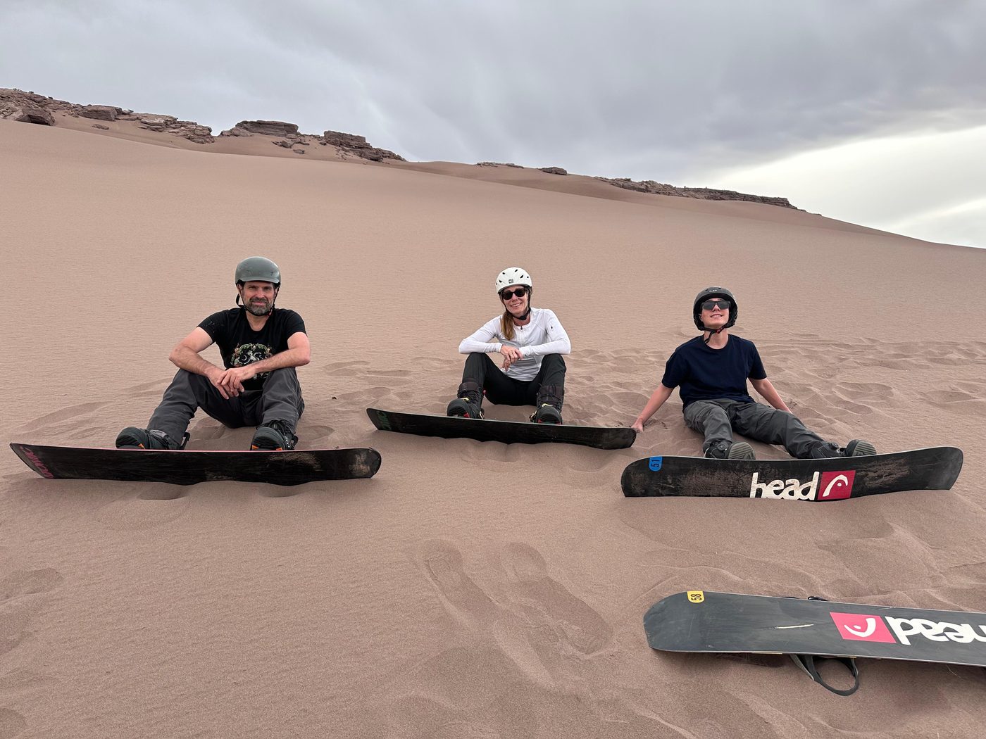 Tres personas de pie sobre sus tablas de sandboard en la cima de una duna, cielo con nubes de atardecer